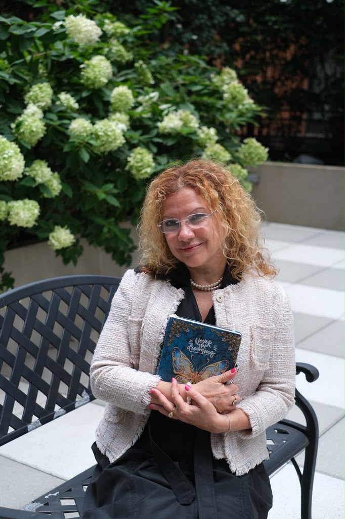 Irina Devi seated on a garden bench holding a notebook, hydrangeas behind her