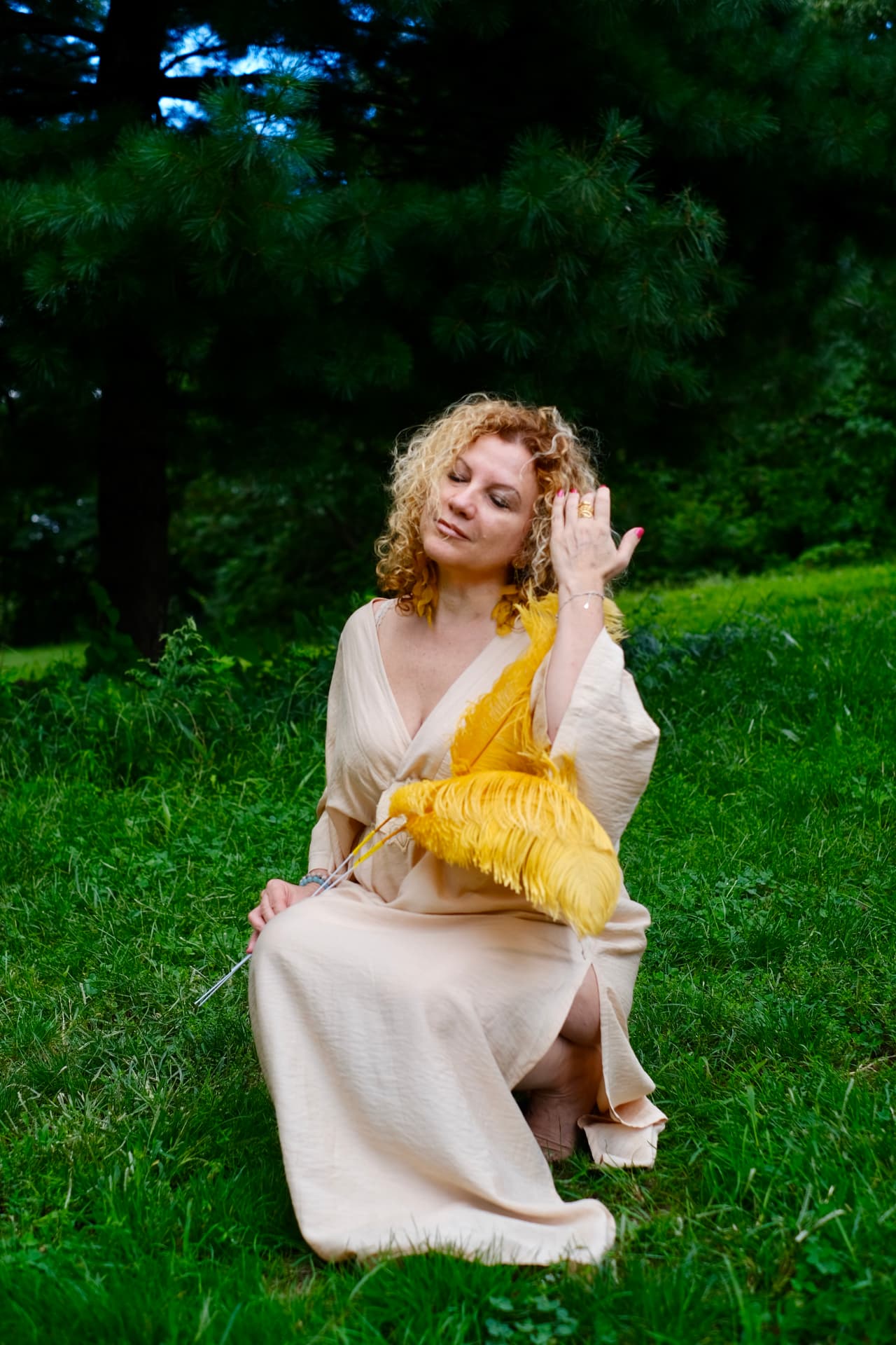 Irina seated on the grass in a cream gown holding golden feathers